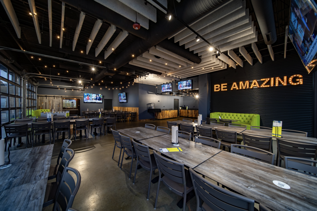 A spacious, modern restaurant with wooden tables, gray chairs, string lights, and multiple TVs. A neon sign on the wall reads BE AMAZING above a green bench. The atmosphere is open and inviting.